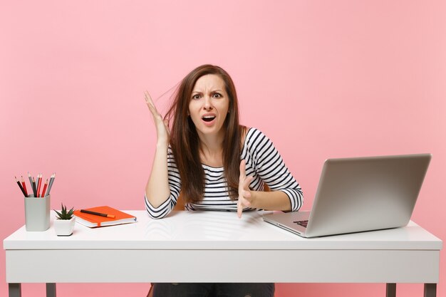 Young exhausted irritated woman in perplexity spreading hands sit, work at white desk with contemporary pc laptop isolated on pastel pink background. Achievement business career concept. Copy space.