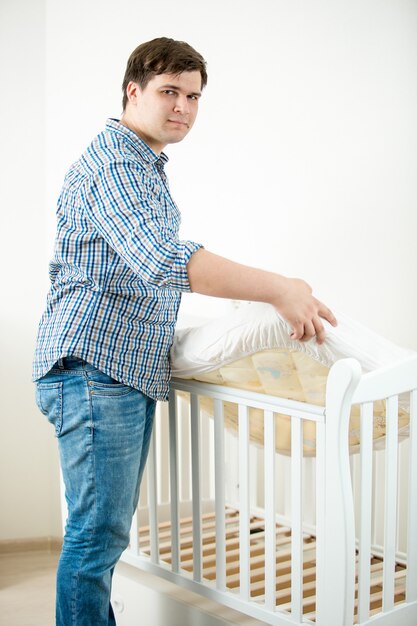 Young expectant father putting mattress in baby's cot at nursery