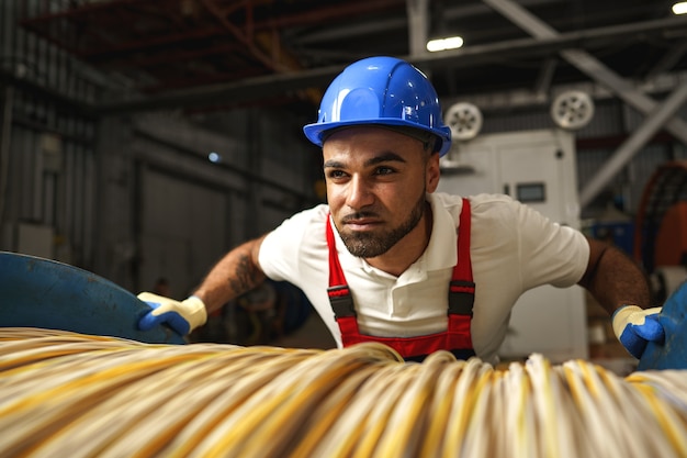 A young factory workman rolls heavy coil of electric cable