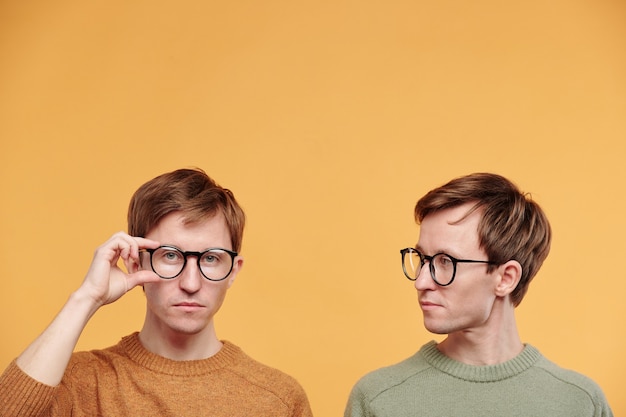 Young fair-haired man in olive sweater looking at brother adjusting glasses against orange background