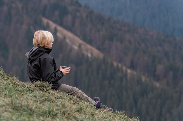 Giovane ragazzo dai capelli biondi si siede su una collina con una tazza di tè ritratto di bambino in montagna viaggi escursioni all'aperto