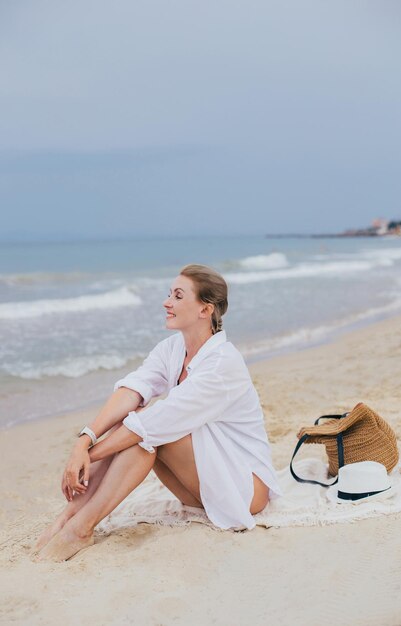 a young fairskinned woman in a white shirt sits alone on the coast against the background of the sea