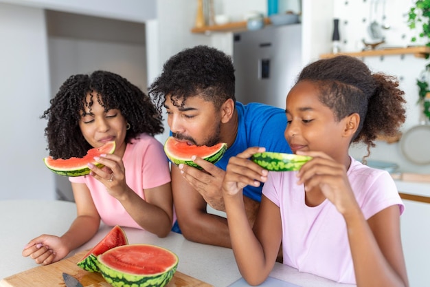 Photo young family eating watermelon and having fun mixed race family in kitchen together eating a watermelon slice