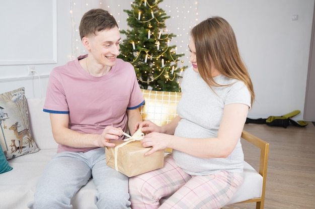 Young family expecting child baby celebrating christmas, opening a gift box. Christmas interior.