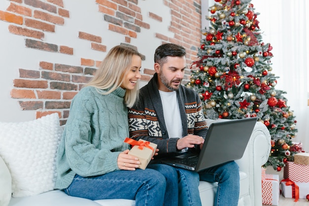 Young family husband and wife choose together Christmas gifts in an online store, sitting at home near the Christmas tree with a laptop