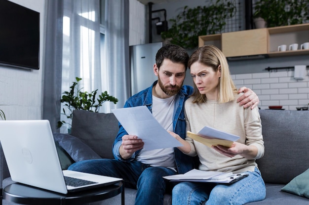 Young family husband and wife reviewing their bills loan and mortgage agreements sitting on the couch at home working with documents on a laptop embarrassed upset