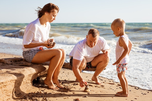 若い家族は、日当たりの良い夏に海のそばで幼い息子と一緒に砂の上で幸せな両親を描いています...