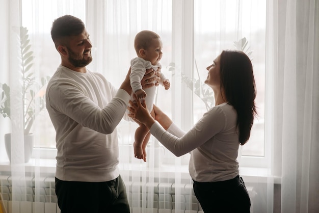 A young family is standing in front of a window, the mother is holding a baby.