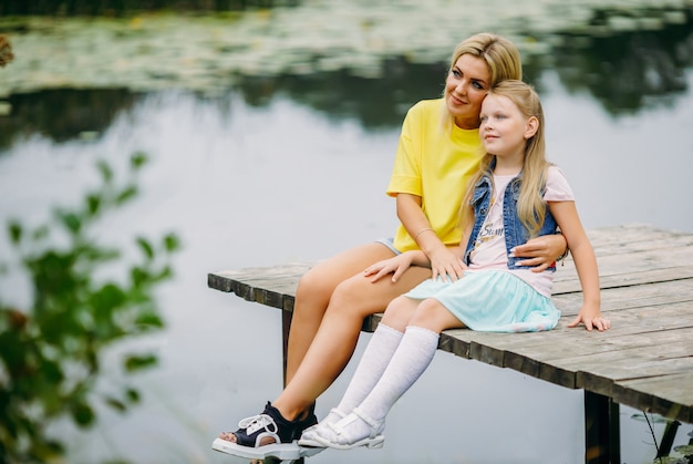 A young family, mother and daughter are sitting on a wooden pier hugging each other and smiling to the side