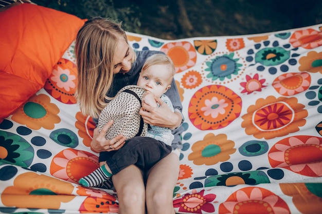 Young family portrait of a beautiful hipster mother with cute blonde boy with blue eyes and vintage hat sitting on a boho hammock outdoors family camping vacation concept