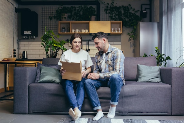 A young family a pregnant woman and a man accidentally received
someone else's parcel they opened the box disappointed sitting at
home on the couch sad the man holds his head