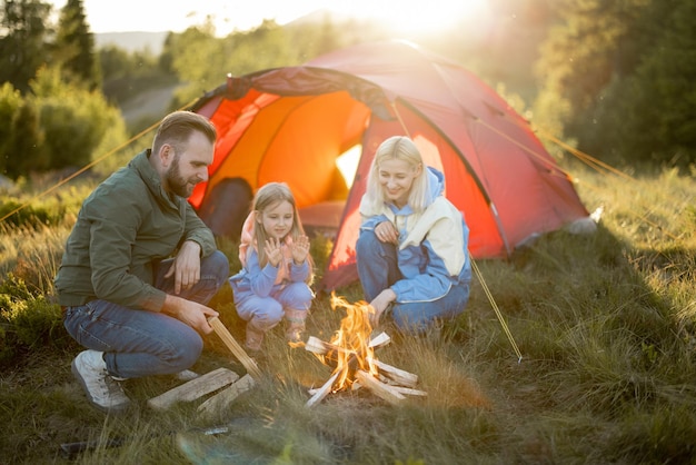若い家族は自然のテントの近くのキャンプファイヤーのそばに座っています