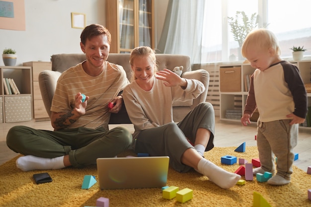 Giovane famiglia seduta sul pavimento e parlando online utilizzando il computer portatile con il loro bambino che gioca vicino a loro a casa