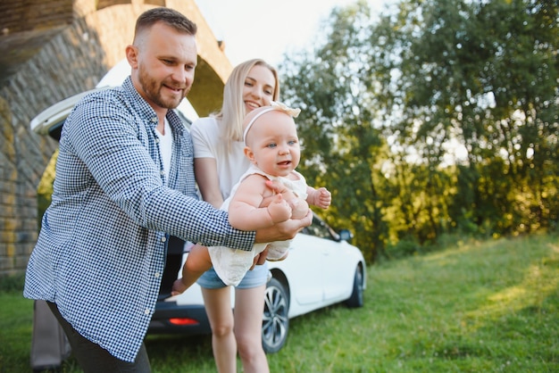 Молодая семья из трех человек в белых одеждах на пикник. Красивые родители и дочь путешествуют на машине во время летних каникул. Сцена в парке