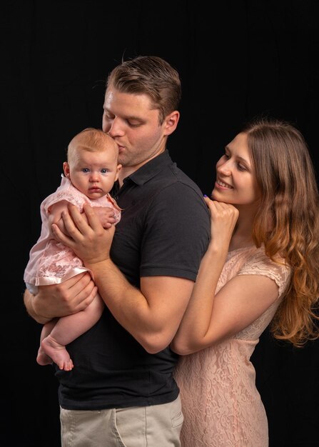 A young family with a baby in their arms Dad kisses the newborn mom looks tenderly The girl is dressed in light clothes the guy is in a black Tshirt on a black background Studio shooting