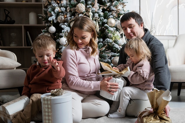 어린 두 아이를 둔 젊은 가족이 휴일에 크리스마스 트리에서 선물 포장을 풀고 있습니다. 크리스마스와 새해의 기쁨과 행복.