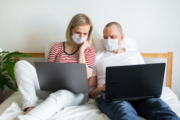 young family works remotely from home on bed at the computer. Quarantined couple coronavirus in medical masks. Stay home safe. Distance learning, education and work. Order food products online