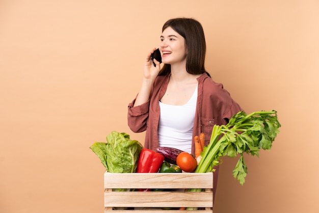 Молодой фермер девушка со свежесобранными овощами в коробке ведет разговор с мобильным телефоном