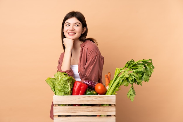 腰に腕を持つポーズと笑顔のボックスで摘みたての野菜を持つ若い農家の少女