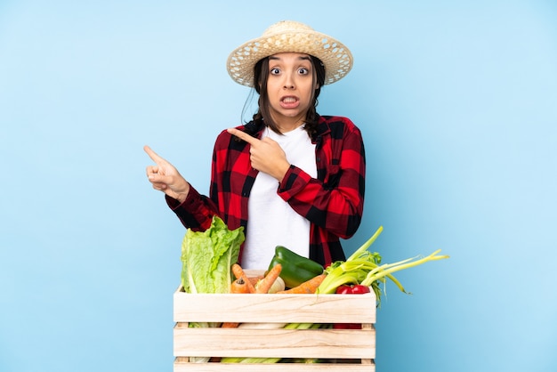 Young farmer Woman holding fresh vegetables in a wooden basket frightened and pointing to the side