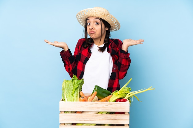 Photo young farmer woman holding fresh vegetables in a wooden basket making doubts gesture