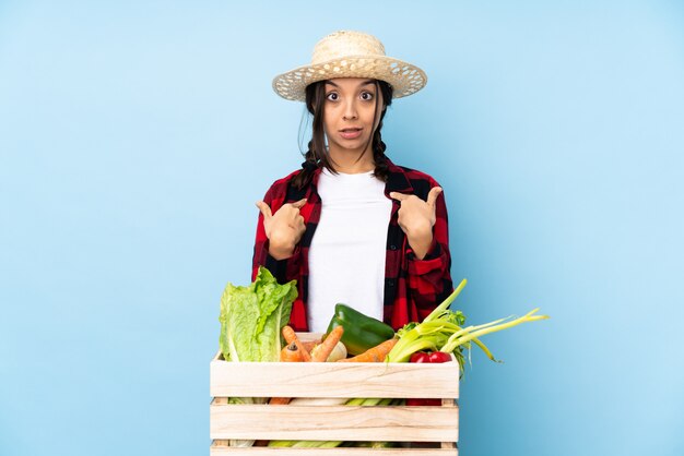 Young farmer woman holding fresh vegetables in a wooden basket with surprise facial expression