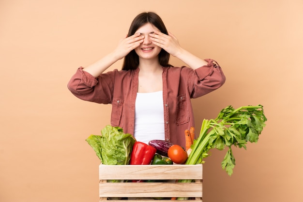 手で目を覆っているボックスで採れたての野菜を持つ若い農家の女性