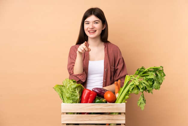 young farmer woman with freshly picked vegetables in a box surprised and pointing front