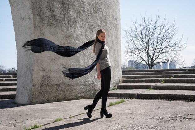 Foto ragazza bionda di giovane modo che porta sciarpa nera lunga sui precedenti della costruzione futuristica.