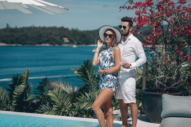 Young fashion couple in sunglasses standing near infinity\
swimming pool on sunny summer day.trip to warm destination.\
honeymoon. beautiful sea landscape. phuket. thailand.