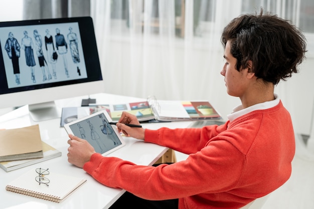 Молодой модельер просматривает новые модели на сенсорной панели, сидя за столом перед экраном компьютера