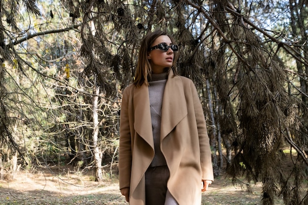 Young fashionable and beautiful girl in a beige coat holding take away tea or coffee and stands alone in the forest