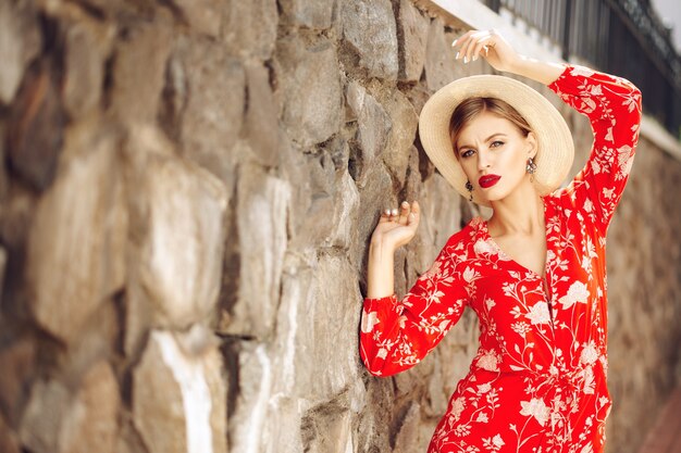 Young fashionable beautiful sexy girl in a red suit and hat stands near a brick wall