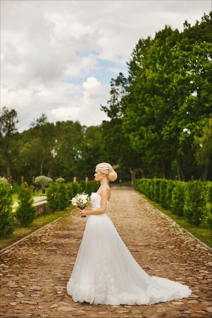 Young fashionable bride, beautiful blonde model girl with stylish wedding hairstyle, in white lace dress posing outdoors with bouquet of flowers in her hands