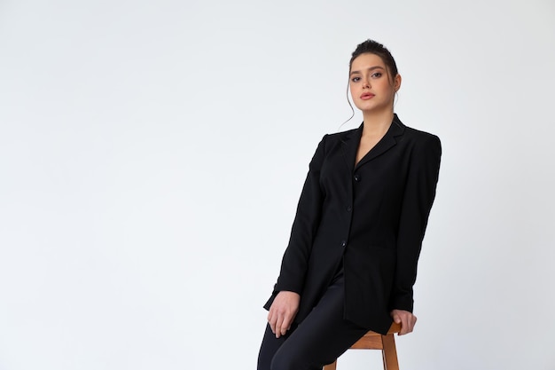 Young fashionable business woman sitting high bar wooden chair dressed black office suit isolated in white background