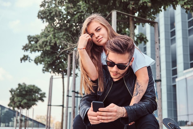 Young fashionable couple relaxing together near skyscraper - pretty girl cuddling her boyfriend while he using smartphone.