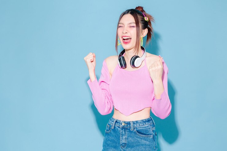 Photo 격리된 파란색 배경에서 축하하는 완벽한 날씬한 몸매를 가진 헤드폰과 롤러 스케이트를 입은 젊고 패셔너블한 귀여운 근심 없는 아시아 여성 실내에서 즐거운 시간을 보내는 긍정적인 모델