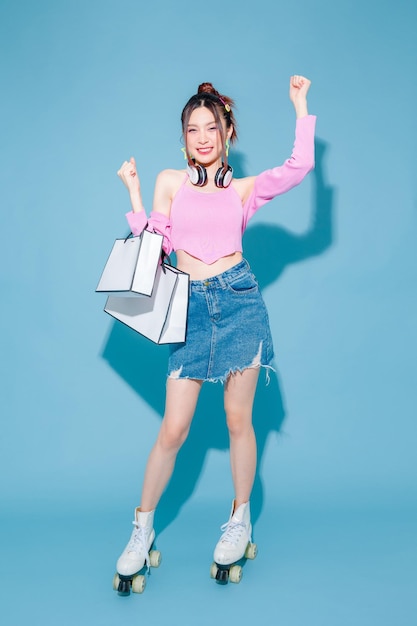 Young fashionable cute carefree Asian woman wearing headphone and roller skate with perfect slim body on isolated blue background Positive model having fun holding shopping bags indoors
