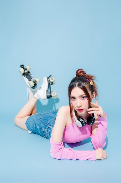 Young fashionable cute carefree asian woman wearing headphone and roller skate with perfect slim body on isolated blue background positive model lying down sexy pose indoors studio