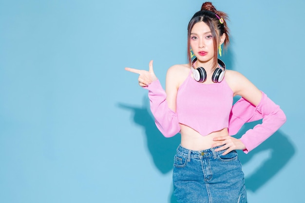 Young fashionable cute carefree Asian woman wearing headphone and roller skate with perfect slim body Pointing to copy space on isolated blue background Female model posing indoors studio