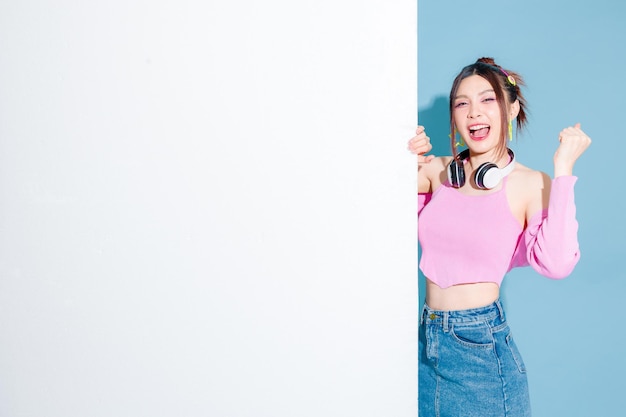 Young fashionable cute successful Asian woman wearing headphone with perfect slim body celebrating peek out blank banner on isolated blue background Positive model having fun indoors