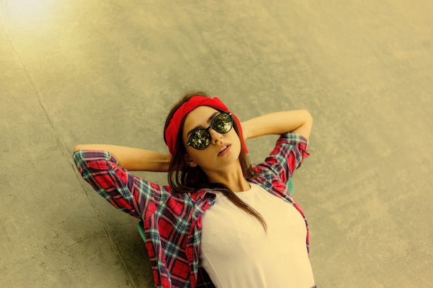 Young fashionable girl in stylish mirrored glasses, red plaid shirt and bandage on her head lies on a concrete surface in skatepark