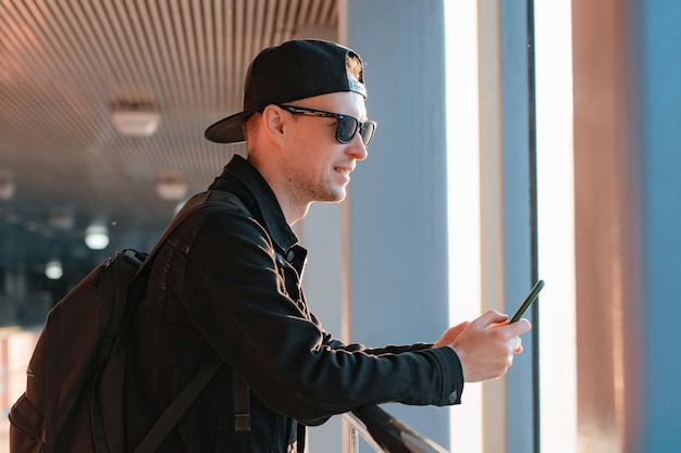 Giovane ragazzo moderno alla moda, un uomo con un berretto nero e occhiali da sole con uno smartphone, parla al telefono su uno sfondo urbano della città in un tunnel sotto i raggi del sole del tramonto.