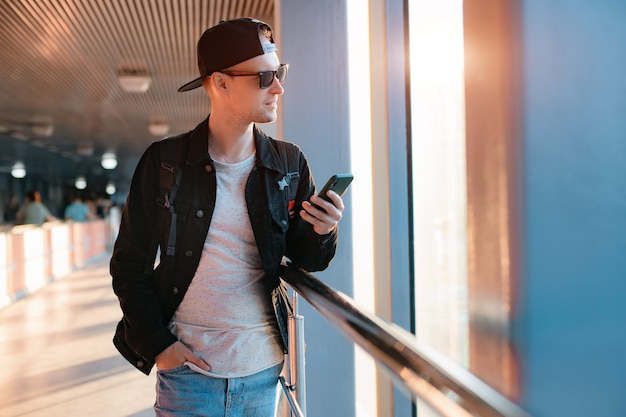 Young fashionable modern guy, a man in a black cap and sunglasses with a smartphone, talks on the phone on a city urban background in a tunnel in the sunset rays of the sun.