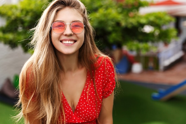 Giovane donna alla moda con capelli biondi che ride e sorride, in piedi nel parco verde in una giornata di sole