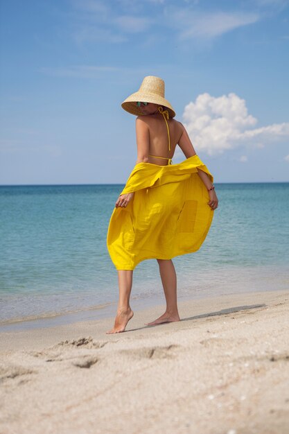 노란색 모자 드레스와 선글라스를 쓴 젊은 유행 여성이 여름 휴가철 해변에서 포즈를 취하고 있다