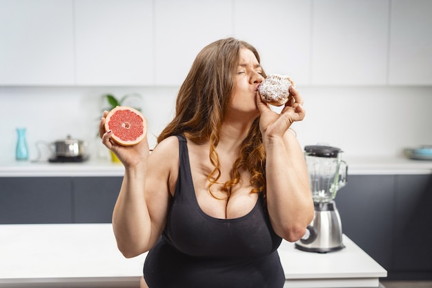 ケーキとグレープフルーツを持って何を食べるかを選ぶ若い太った女性。不健康な食べ物を食べる美しいぽっちゃり若い女性。