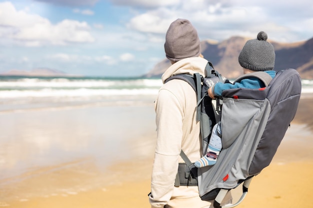 Young father carrying his infant baby boy son in backpack on windy sandy beach Family travel and winter vacation concept