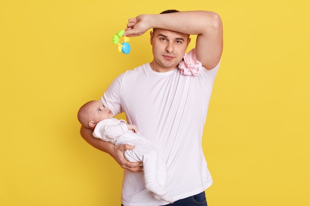 Young father dad frustrated at crying baby, caucasian male wearing white t shirt keeping hand on forehead, holding kid's toy in hand.