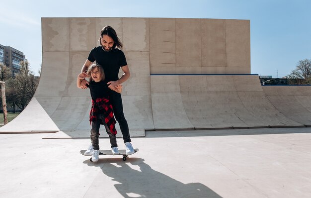 Young father helps his little son to ride on the skateboard in a skate park at the sunny day .